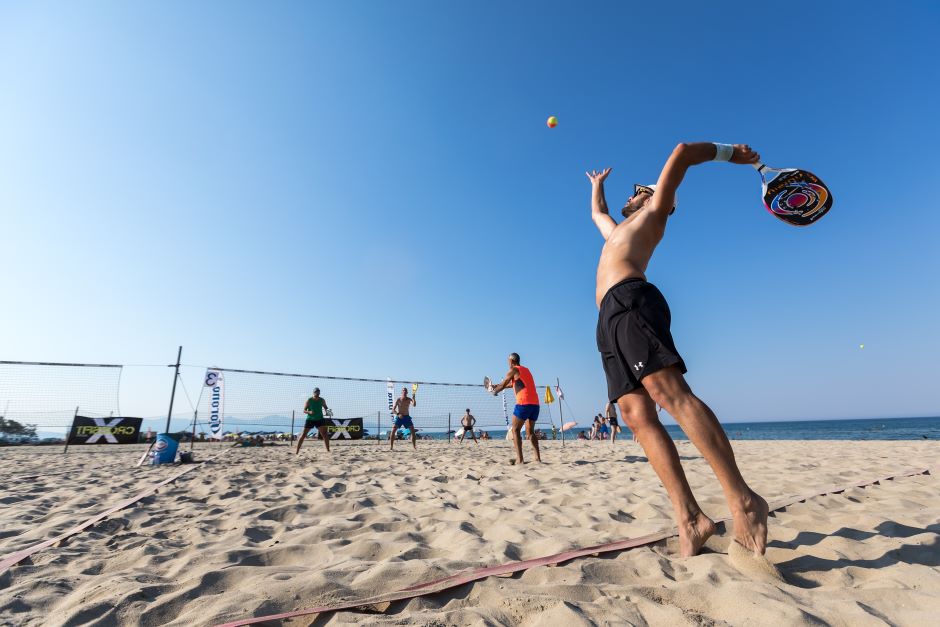 Quatro jogadores homens em uma quadra de areia de beach tennis à beira-mar, um dos jogadores está prestes a realizar um saque