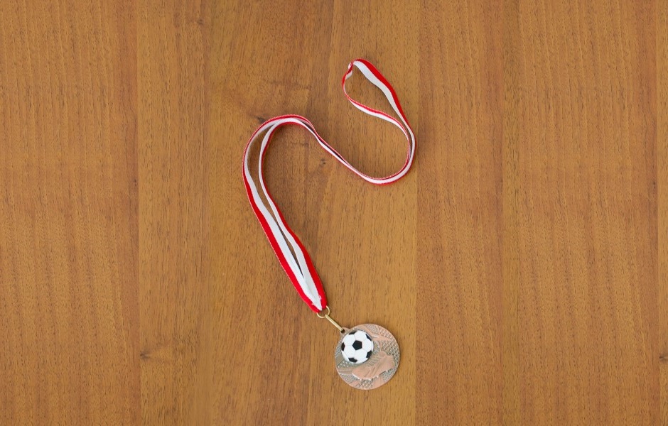 Imagem de uma medalha com um ícone de bola de futebol e uma chuteira gravados em seu interior, disposta sobre uma superfície de madeira.