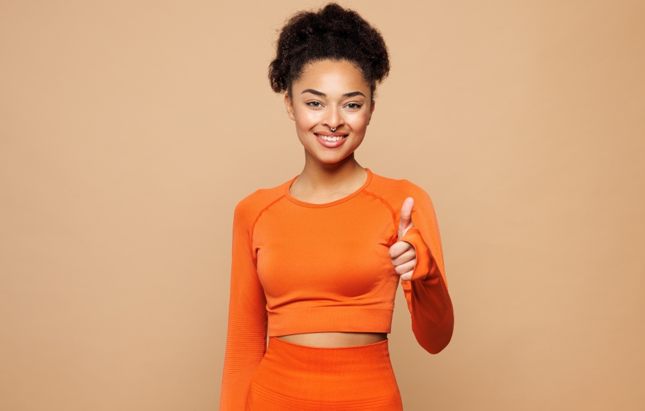 Mulher jovem e sorridente vestida com roupa laranja, mostrando entusiasmo e positividade, em fundo neutro.