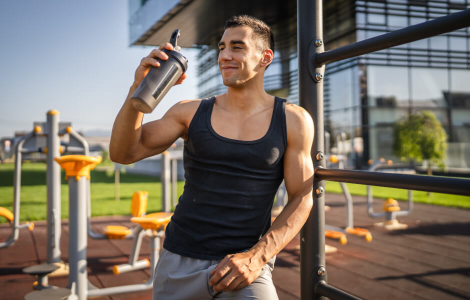 Foto de um homem em uma academia ao ar livre segurando um shake de suplemento.