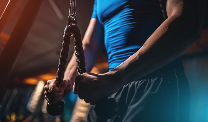 Homem com camiseta azul treinando força com corda de resistência na academia, focado e determinado na atividade de exercícios físicos para hipertrofia muscular.