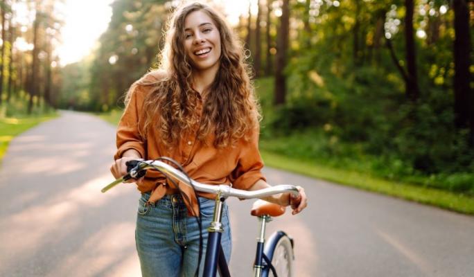 Jovem mulher sorridente com cabelo encaracolado, usando blusa laranja e jeans, segurando bicicleta em uma estrada asfaltada cercada por árvores verdes em um dia ensolarado.