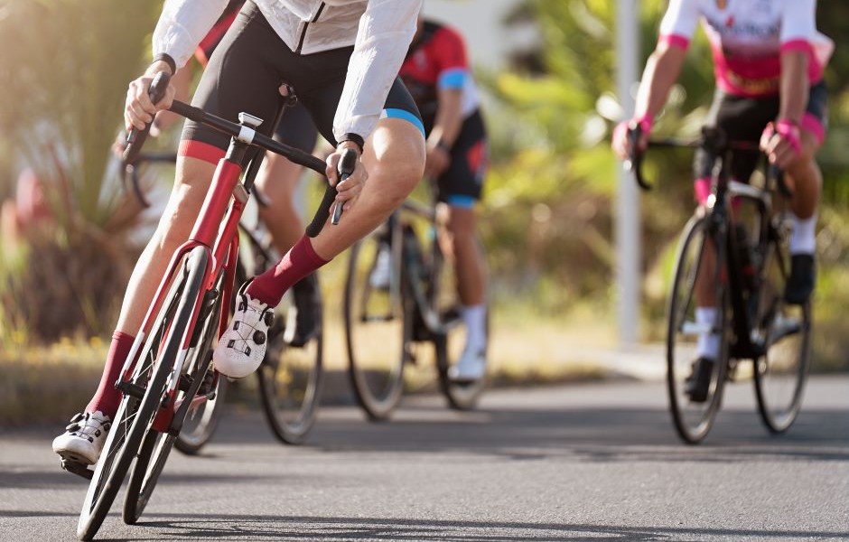 Grupo de ciclistas participando de uma corrida em um circuito ao ar livre, com destaque para um ciclista de cima de uma bicicleta de corrida, vestindo roupas esportivas coloridas e capacete, no ambiente ao ar livre com vegetação ao fundo.