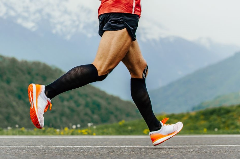 Imagem mostra as pernas de um homem correndo. É possível ver suas meias e tênis de corrida. Ao fundo podem-se ver montanhas.