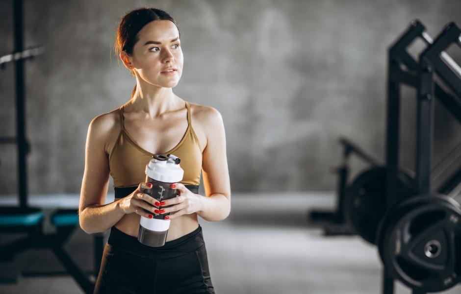 Mulher em trajes esportivos segurando uma coqueteleira de suplemento dentro de uma academia.