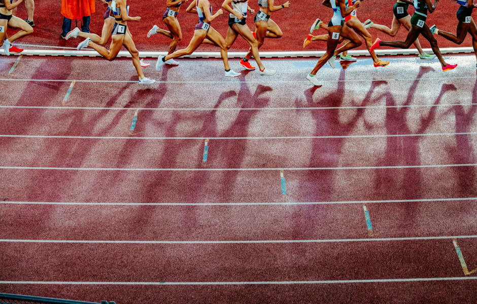 Imagem de uma pista de atletismo com vários corredores em ação, destacando suas pernas em movimento.