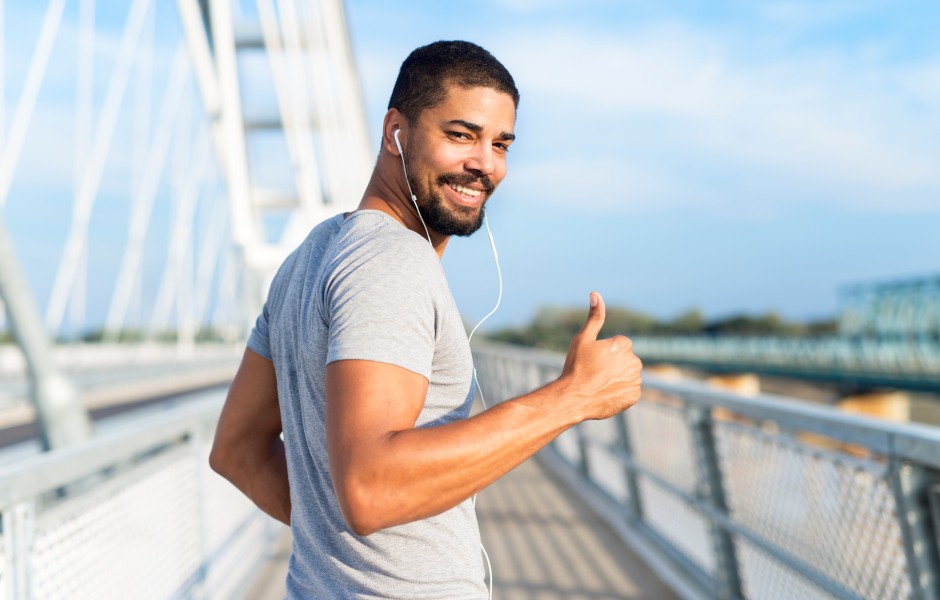 Homem jovem e sorridente com barba, usando camiseta cinza e fones de ouvido brancos, corre sobre uma ponte metálica em um dia ensolarado. Ele olha para a câmera e faz um sinal de positivo com o polegar. O fundo mostra a estrutura da ponte e o céu azul.