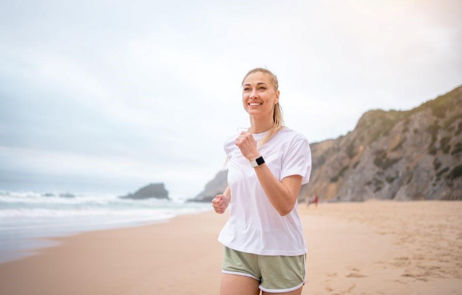 Mulher praticando corrida na praia, sorrindo e aproveitando o exercício ao ar livre com o mar ao fundo e uma paisagem de falésias ao longe.