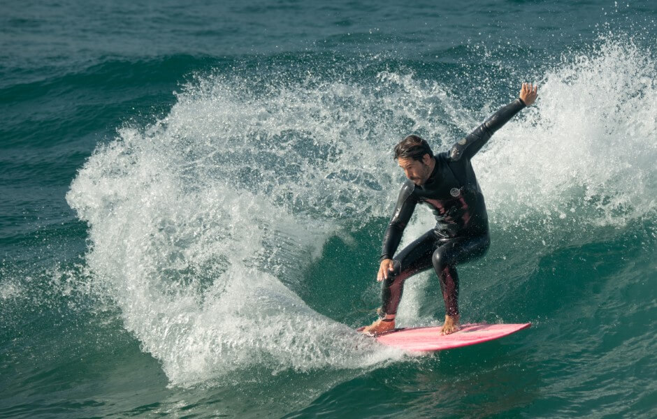 Imagem de um surfista pegando uma onda, ele está em pé sobre a prancha e usa uma roupa de borracha.