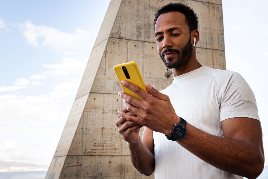 Imagem de um homem mexendo em um celular que possui a capa amarela. O homem também usa um smartwatch em seu punho esquerdo e fones de ouvido sem fio. O homem parece estar em cima de uma ponte.