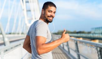 Homem jovem e sorridente com barba, usando camiseta cinza e fones de ouvido brancos, corre sobre uma ponte metálica em um dia ensolarado. Ele olha para a câmera e faz um sinal de positivo com o polegar. O fundo mostra a estrutura da ponte e o céu azul.