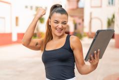 Mulher jovem comemora vibrante ao olhar para um tablet em ambiente externo. A imagem expressa sucesso, conquista de metas e felicidade com resultados digitais.