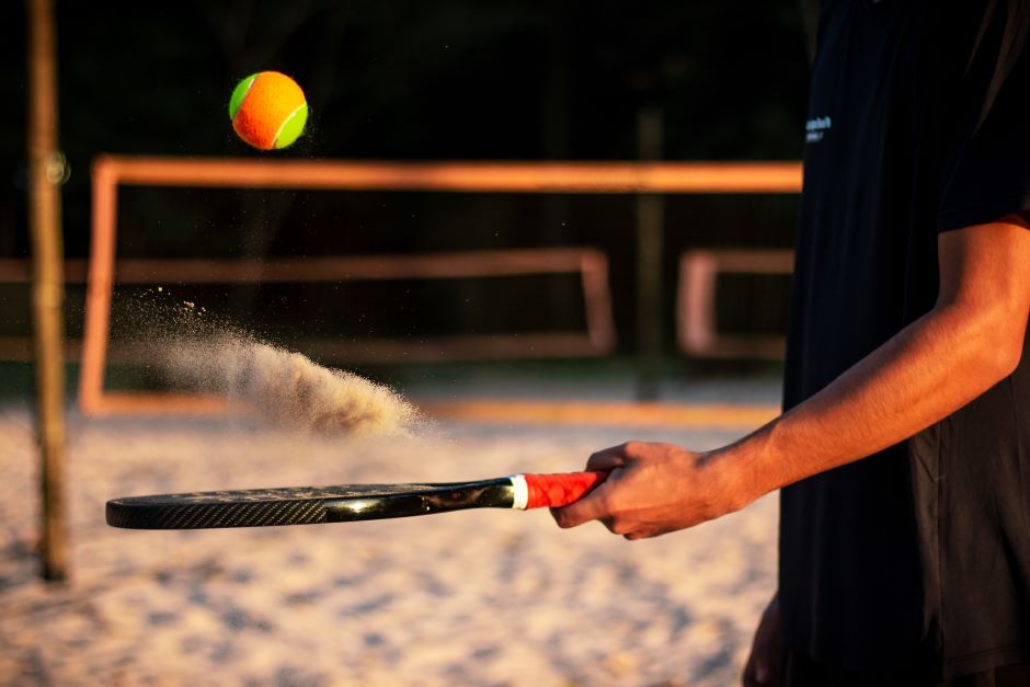 Atleta segurando raquete de beach tennis com bola de beach tennis voando sobre a raquete, fundo com redes e areia de uma quadra de beach tennis