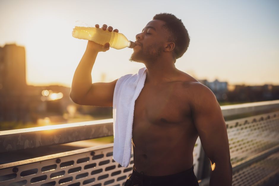 Imagem de um homem, sem camiseta e com uma toalha estendida em seu braço direito, ele está bebendo água em uma garrafa após prática de atividade física, o homem está em uma ponte, fundo da imagem mostrando uma cidade em um dia de céu ensolarado