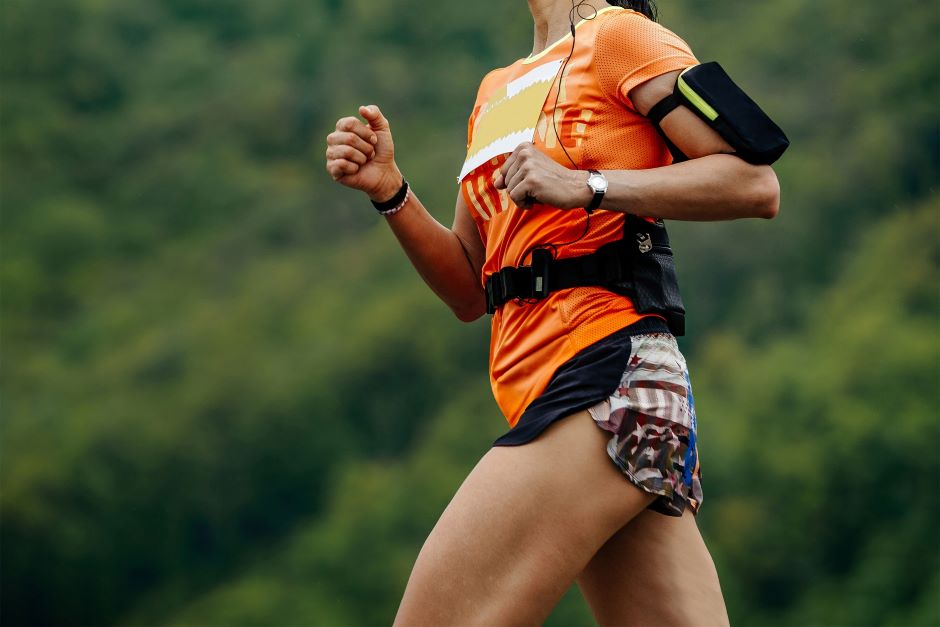 Imagem de uma mulher correndo e utilizando acessórios como uma braçadeira e fones de ouvido com fio, além de roupas específicas para a prática de corrida. Fundo da imagem borrado, parecem ser árvores