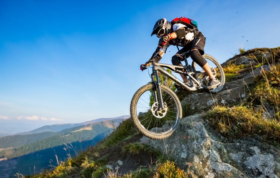 Mountain biker descendo uma trilha íngreme de pedra e terra em meio a uma paisagem de montanhas e céu claro, praticando mountain biking de alta altitude.