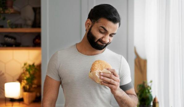 Homem jovem com barba cheia e cabelo escuro, vestindo camiseta cinza justa, em pé dentro de uma cozinha. Ele está olhando para baixo com expressão de dúvida ou hesitação enquanto segura um hambúrguer com gergelim envolto em um papel pardo.