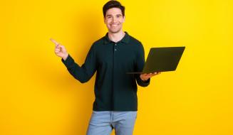 Promotor de eventos sorridente segurando um laptop em frente a um fundo amarelo. Ele aponta para a direita, transmitindo energia positiva e entusiasmo. Ideal para temas de tecnologia e produtividade.