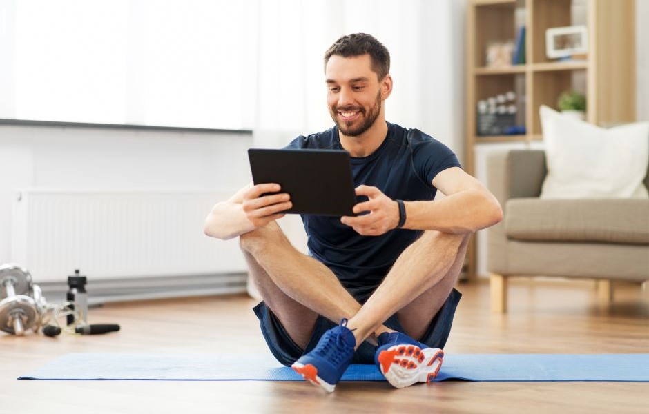 Homem sorridente exercitando-se em casa, sentado no tapete de yoga enquanto assiste algo em um tablet, ambiente acolhedor com móveis e equipamentos de ginástica ao fundo.