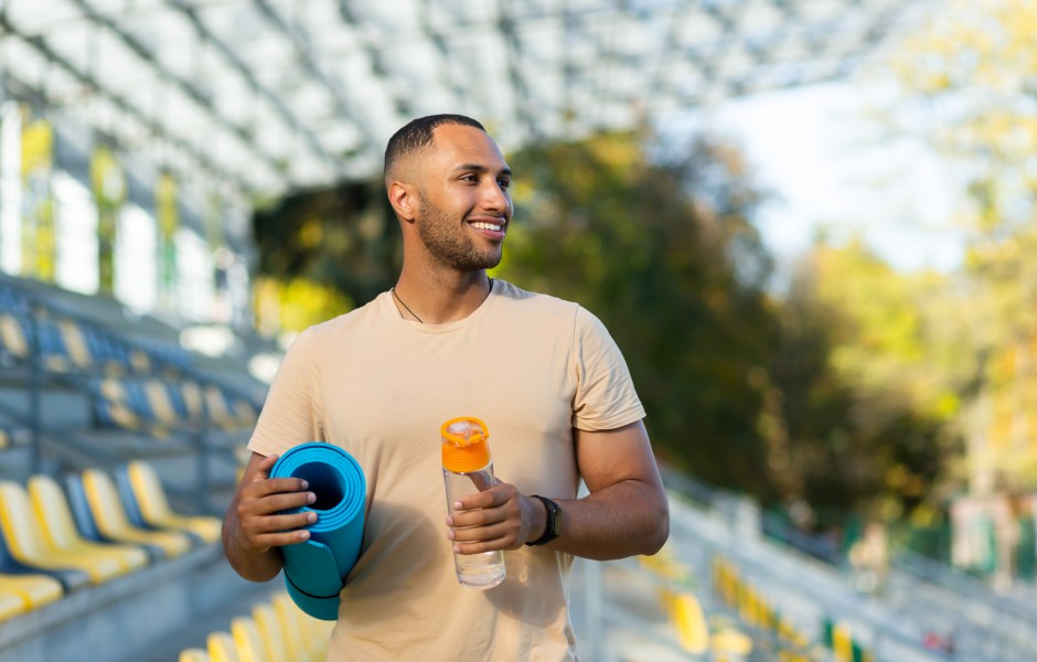  Um jovem atleta sorridente caminha por um estádio carregando um tapete de yoga azul e uma garrafa de água.