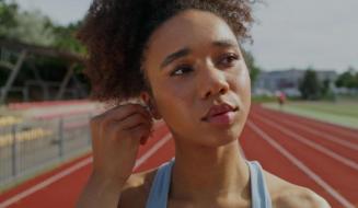 Mulher jovem negra com cabelo cacheado ajustando fone de ouvido sem fio enquanto se prepara para treinar em uma pista de atletismo vermelha ao ar livre.