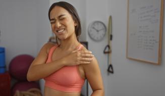 Imagem de uma mulher com dor no ombro. Ela usa um top esportivo rosa e mostra expressão de desconforto, indicando lesão.