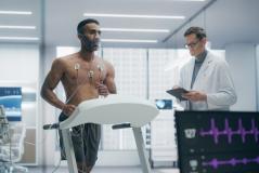 Paciente masculino realizando testes cardíacos com eletrodos no peito sob supervisão de um médico em laboratório de medicina esportiva.