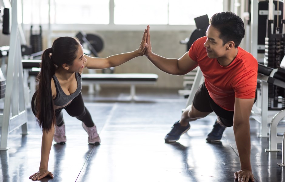 A imagem mostra um homem e uma mulher se exercitando em uma academia. Eles estão em posição de prancha, com os corpos voltados para baixo, apoiados nos pés e em uma das mãos. Com a outra mão, eles se estendem e seguram as mãos um do outro.