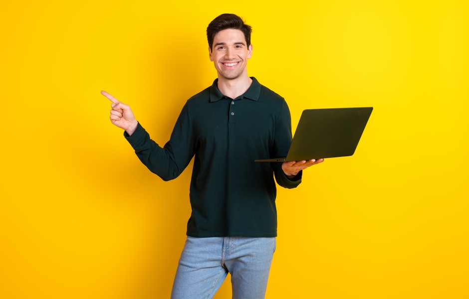Promotor de eventos sorridente segurando um laptop em frente a um fundo amarelo. Ele aponta para a direita, transmitindo energia positiva e entusiasmo. Ideal para temas de tecnologia e produtividade.
