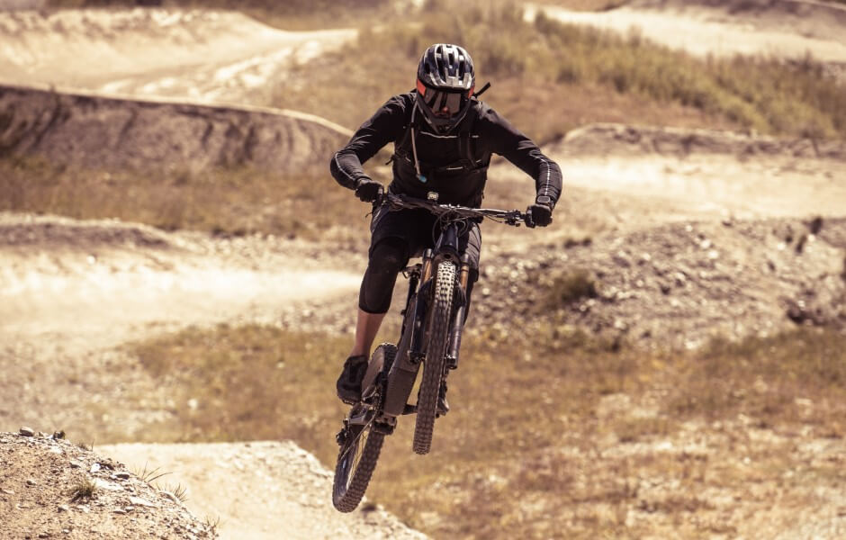 Imagem de um praticante de mountain bike no ar, em meio a um salto sobre uma estrada de terra.