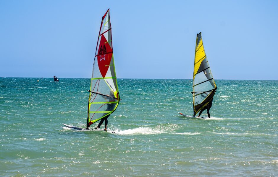 Imagem com dois praticantes de windsurf em destaque. Eles deslizam pelo mar azul sob pranchas com velas acopladas.