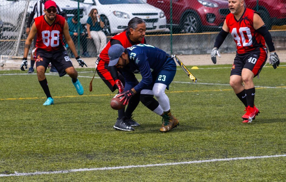 Imagem de quatro atletas de flag football em um gramado. Os jogadores têm fitas presas às cinturas; um jogador segura uma bola semelhante à de futebol americano, enquanto outro tenta tirar a fita de sua cintura.