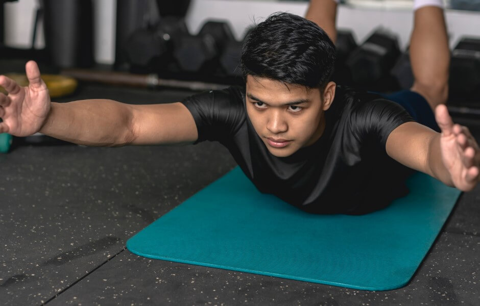 Imagem de um rapaz executando o exercício "Superman" em um ambiente de academia, olhando concentrado para frente.