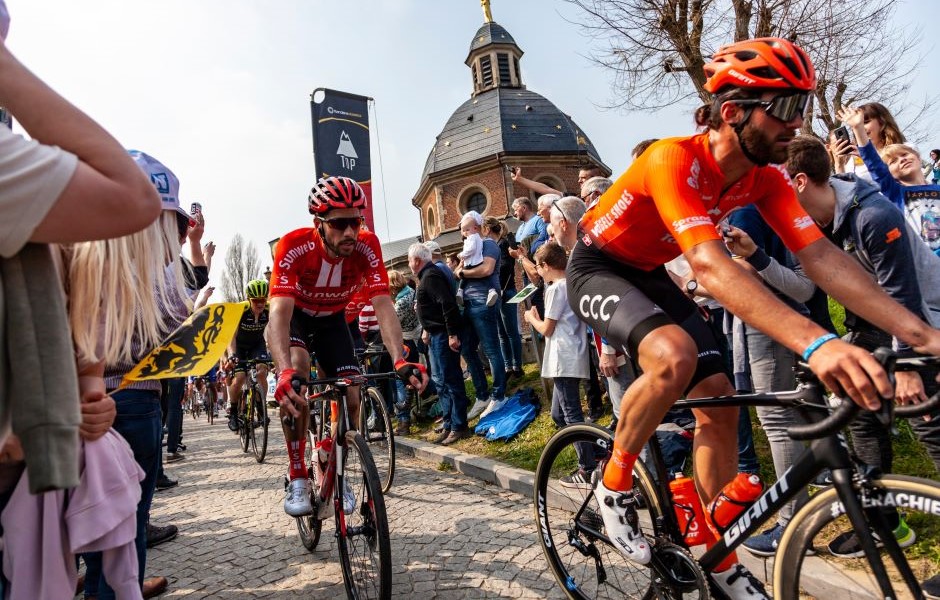 Atletas profissionais de ciclismo competindo em uma corrida oficial com uma multidão de espectadores apoiando ao lado de uma igreja histórica ao ar livre.
