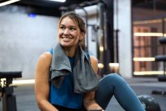Mulher sorridente no supermercado com roupas esportivas, segurando um celular, após treino ou atividade física, ambiente moderno e bem iluminado.
