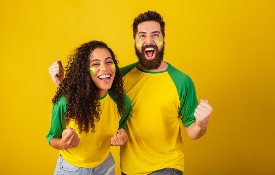 A imagem mostra dois torcedores em um fundo amarelo vibrante. Eles estão vestidos com camisetas de cores que remetem à seleção brasileira. O homem à direita, com barba e cabelo escuro, grita alegremente com punhos cerrados. A mulher à esquerda, de cabelos cacheados, sorri e celebra.