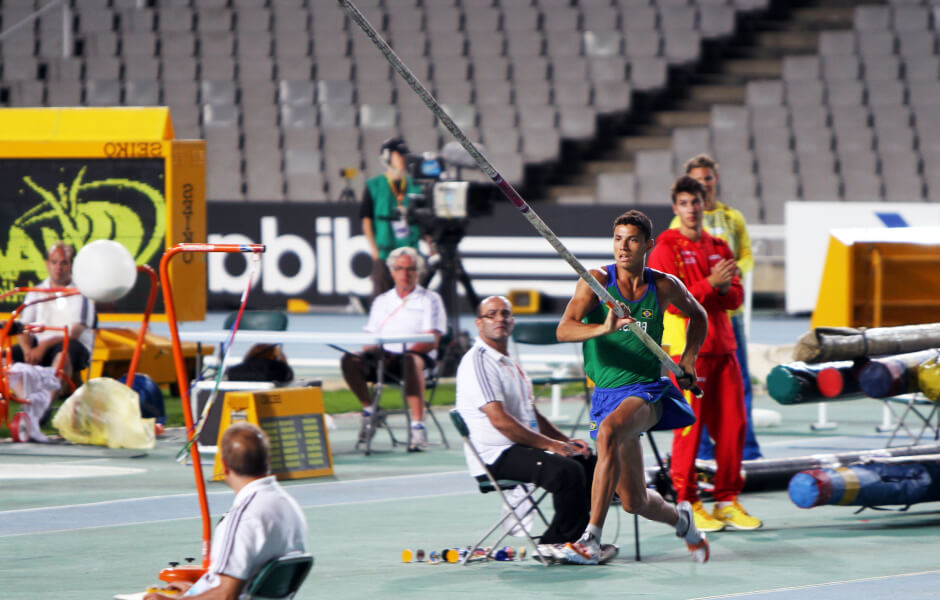 Imagem de um atleta em movimento, usando uniforme da seleção brasileira e segurando uma vara de salto com vara.