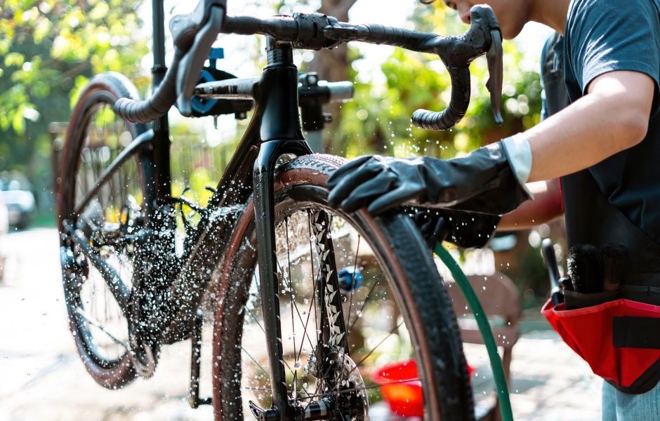 Pessoa lavando uma bicicleta ao ar livre com água, usando luvas de proteção, em um ambiente ensolarado com árvores ao fundo.