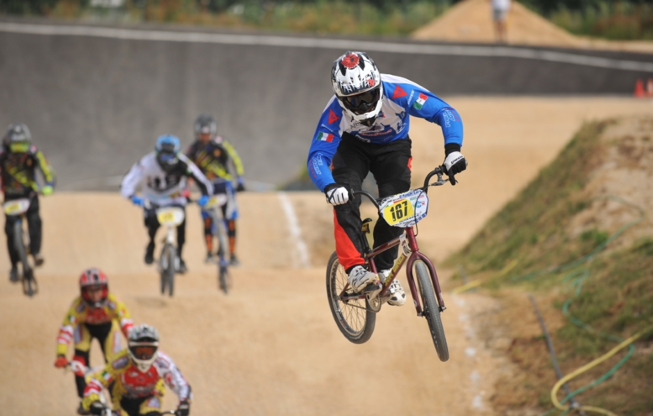 Imagem de um ciclista realizando um salto durante uma competição de BMX. Vários outros ciclistas podem ser vistos ao fundo na pista de corrida. O ciclista em destaque usa um traje azul e um capacete refletivo.