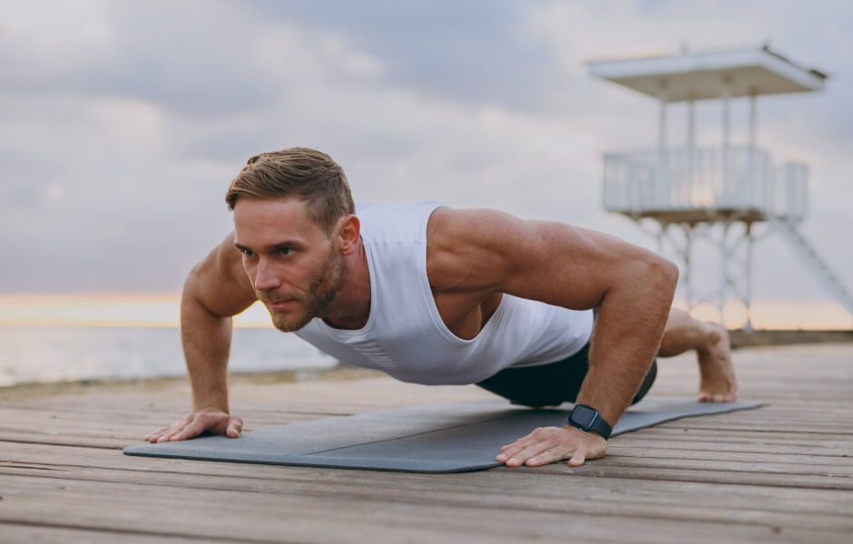 Um homem executando uma flexão de braços sobre uma superfície de madeira próxima ao mar. Ele está usando uma regata e olha concentrado para frente.