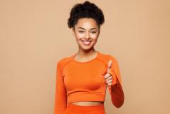 Mulher jovem e sorridente vestida com roupa laranja, mostrando entusiasmo e positividade, em fundo neutro.