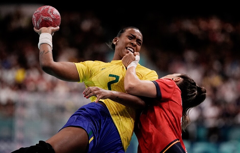 Jogadora de handebol do Brasil em posição de ataque e segurando uma bola de handebol na mão esquerda. A jogadora brasileira está sendo marcada por uma jogadora espanhola.