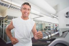 Homem praticando corrida na esteira em uma academia moderna, com sorriso feliz e usando roupas de ginástica brancas.