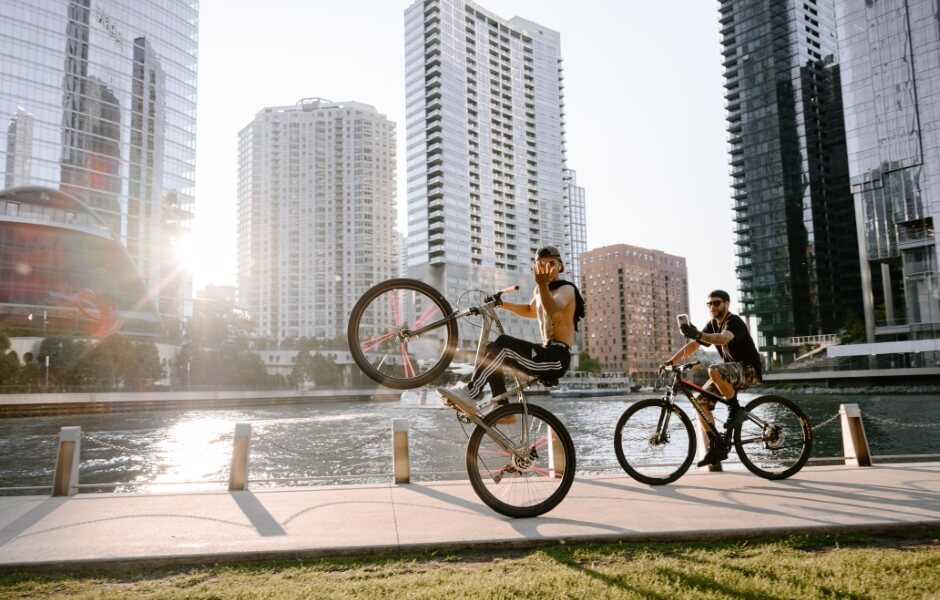 Dois ciclistas realizando manobras em bicicletas modernas no calçadão à beira do rio, com arranha-céus ao fundo durante o pôr do sol.