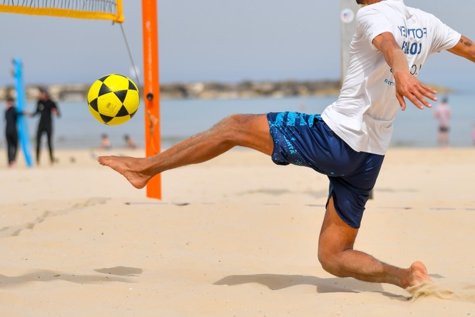 Homem em posição de chute e em direção para bater em bola de futevôlei, quadra de areia e rede de futevôlei no fundo da imagem